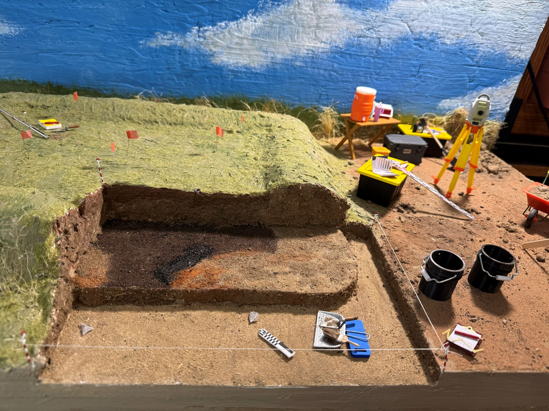 Archaeological dig site model with tools, soil layers, buckets, and measuring equipment.